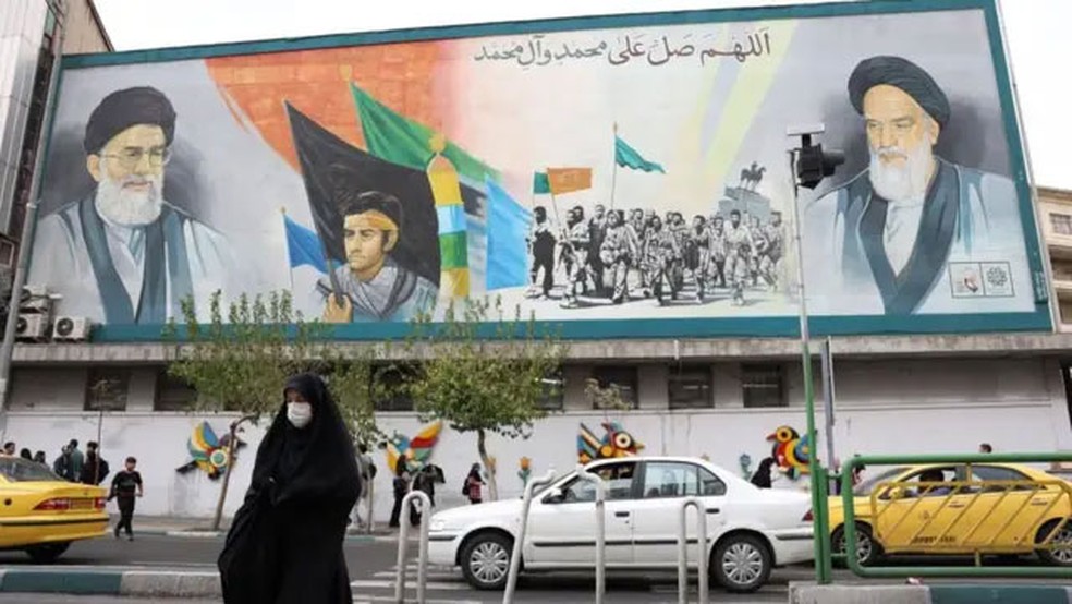 Mulher passa em frente a pintura do líder supremo do Irã aiatolá Ali Khamenei e o antecessor Ruhollah Khomeini em Teerã — Foto: EPA via BBC