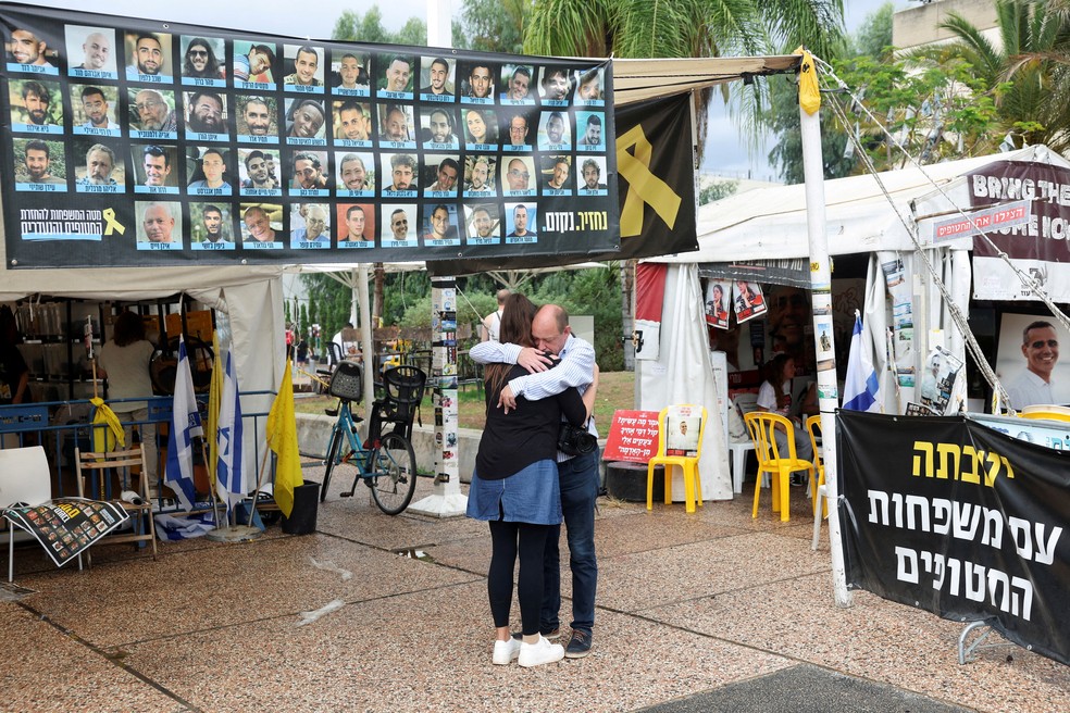 Pessoas se abraçam ao lado de uma faixa com fotos de reféns na "Praça dos Reféns" em Teal Aviv — Foto: REUTERS/Ronen Zvulun