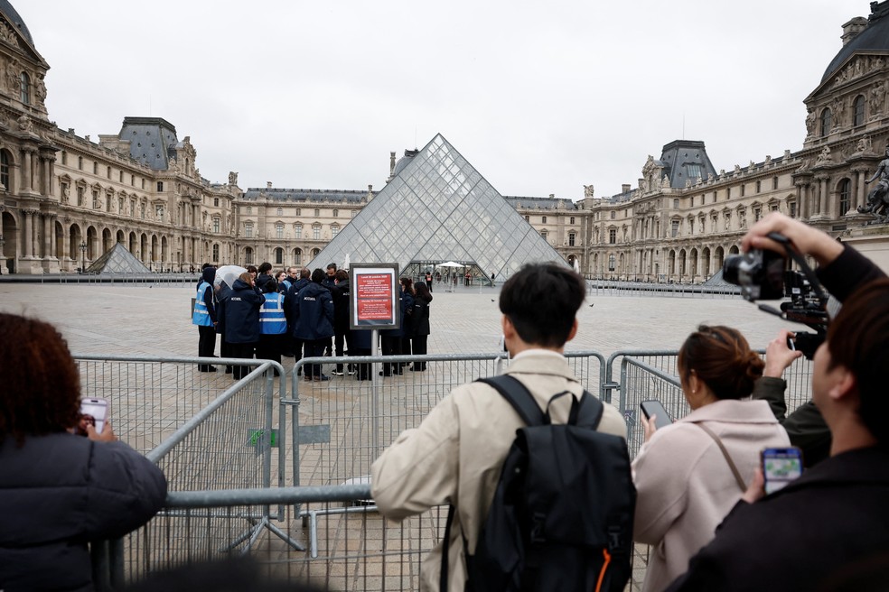 Visitantes perto da Pirâmide de vidro do Louvre observam, por trás de alambrado, placa anunciando que museu permaneceria fechado em 20 de outubro de 2025 após roubo de joias. — Foto: REUTERS/Benoit Tessier