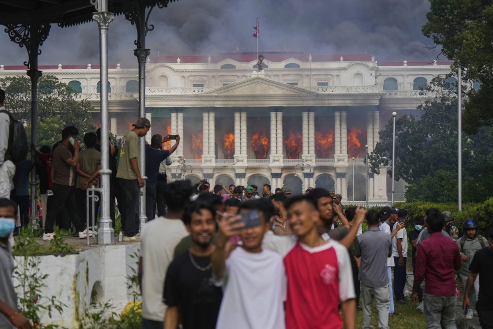 Jovens tiram selfie com o palácio do governo do Nepal em chamas ao fundo, em 9 de setembro de 2025 — Foto: AP Photo/Niranjan Shrestha