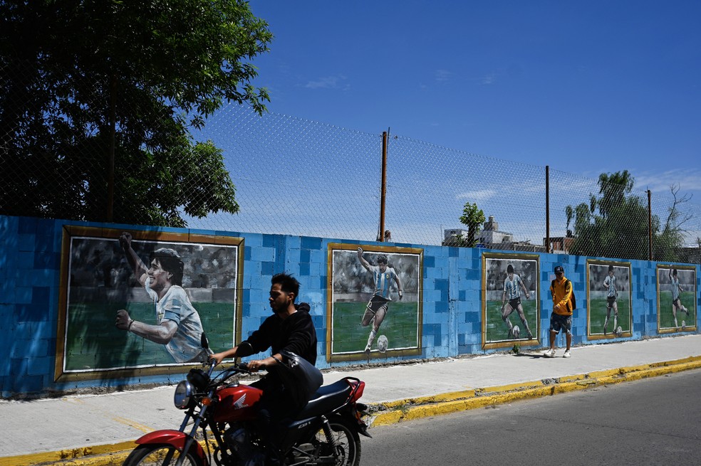Um motociclista e um homem passam por um mural com imagens do ídolo do futebol argentino Diego Armando Maradona em Villa Fiorito, nos arredores de Buenos Aires. — Foto: LUIS ROBAYO/AFP