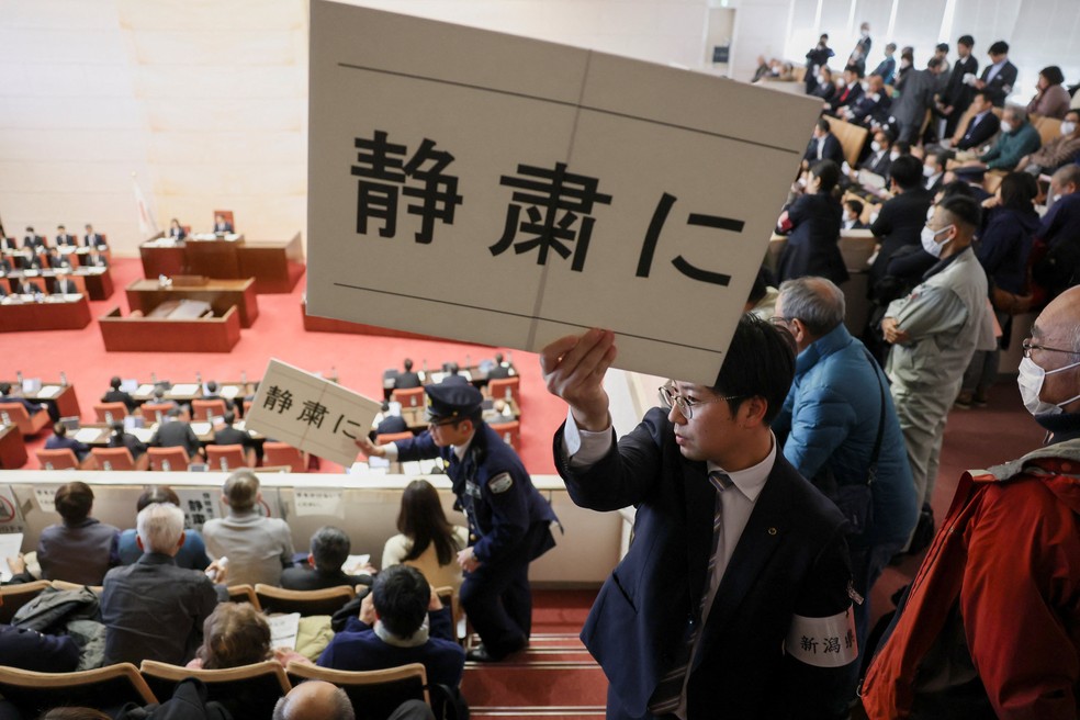 Agentes de segurança seguram cartazes com os dizeres "Silêncio, por favor" perto dos parlamentares da Assembleia da Prefeitura de Niigata, que participam de uma votação de confiança nas decisões do governador da prefeitura sobre a retomada parcial das operações da Usina Nuclear de Kashiwazaki Kariwa, da Tokyo Electric Power Company (TEPCO) — Foto: REUTERS/Issei Kato