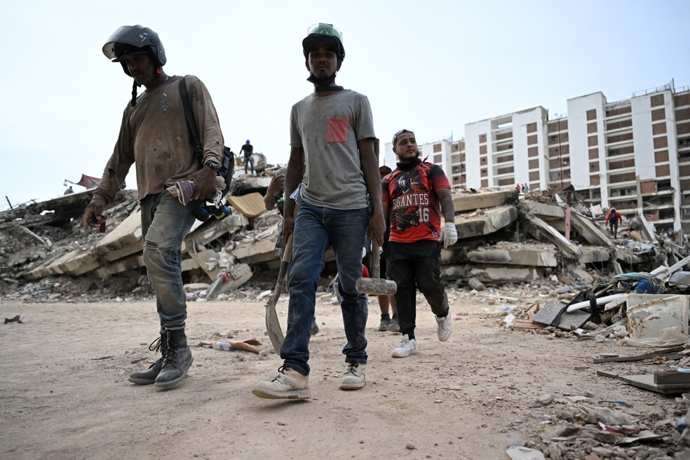 Trabalhadores carregam ferramentas enquanto caminham entre os escombros de prédios desabados em Caraballeda, estado de La Guaira, Venezuela, em 9 de julho de 2026 — Foto: RAUL ARBOLEDA / AFP